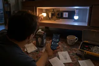 Person crouched under a living room console, comparing two different smart bulbs in a realistic home setting with mixed lighting and installation tools nearby