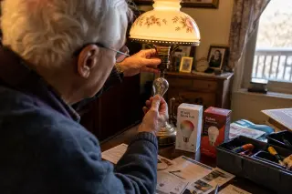 Senior swapping an old bulb from a lamp, with WiFi and Zigbee smart bulbs in packaging on a table in a cozy, softly lit living room