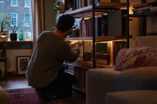 Homeowner installing smart LED strip beneath bookshelf, creating cozy accent lighting in a stylish, eclectic living room