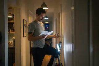 A person on a step stool installing a smart dimmer in a dim hallway of a partly renovated apartment, studying a manual under mixed lighting from smart bulbs and a work lamp.