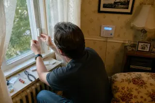 DIY enthusiast installing a glass break sensor on a window in a retro living room, seen from above their shoulder, with tools and an alarm panel visible in natural afternoon light.