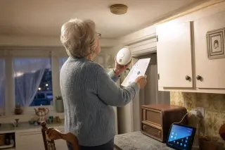 Senior woman comparing a classic and a smart smoke detector in a lived-in kitchen, absorbed in installation under warm evening light.