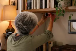 Senior woman adjusting a discreet camera beneath a bookshelf in a cozy, warmly lit living room, integrating technology with home decor