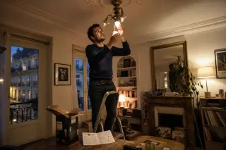 Tenant carefully installs a smart bulb in a vintage ceiling lamp in a Paris apartment, with instruction manual and packaging visible on a nearby table under warm evening light.